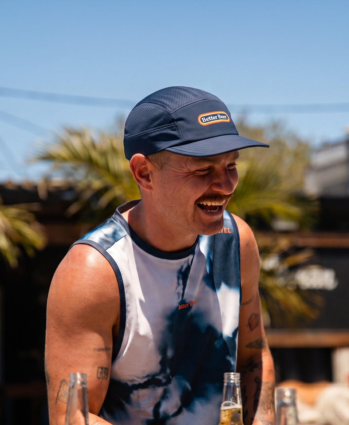 A man outdoors wearing a custom running cap with a visible 'Better Beer' logo and a tie-dye singlet, smiling while holding beer bottles in a sunny outdoor setting.