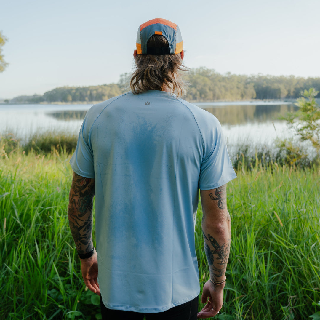 Man wearing a light blue t-shirt and colorful cap standing in a grassy field with a lake in the background